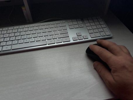 Close-up of a hand using a computer mouse beside a white keyboard on a light-colored desk.